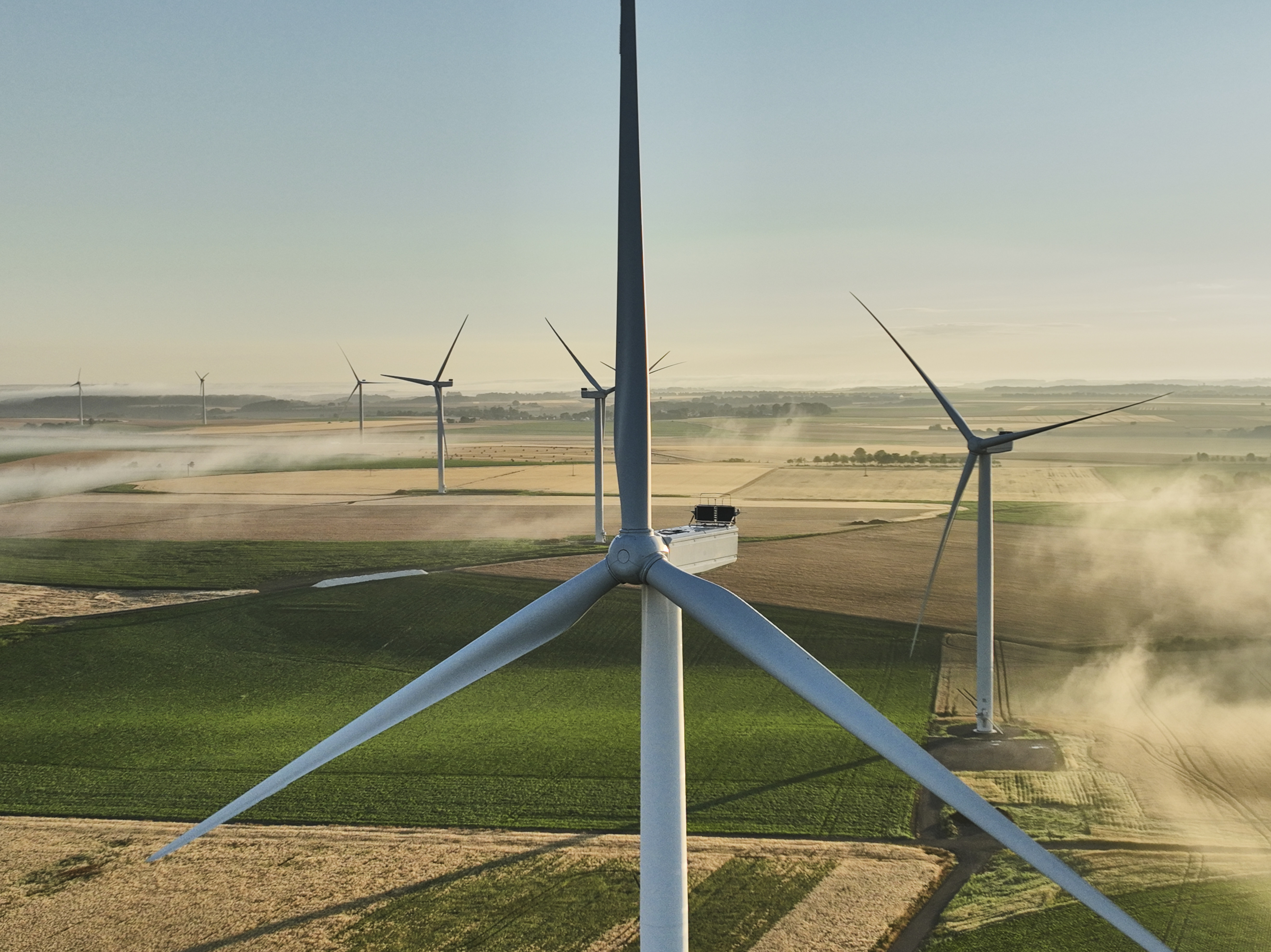 Aerial view of multiple wind turbines on a vast landscape with fields and mist in the early morning light.
