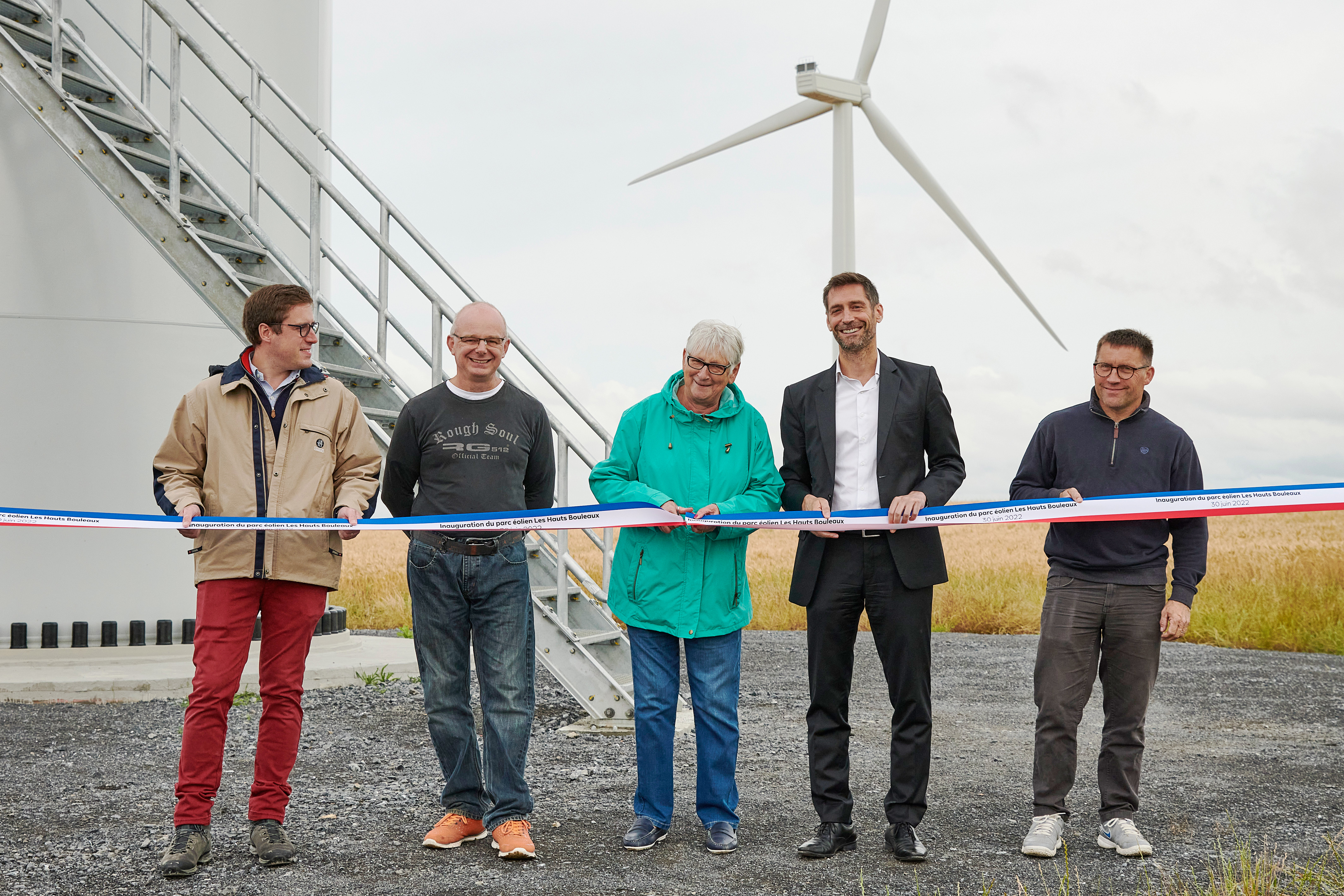 Un groupe de personnes célèbre l'inauguration d'un parc éolien, tenant un ruban près d'une éolienne.