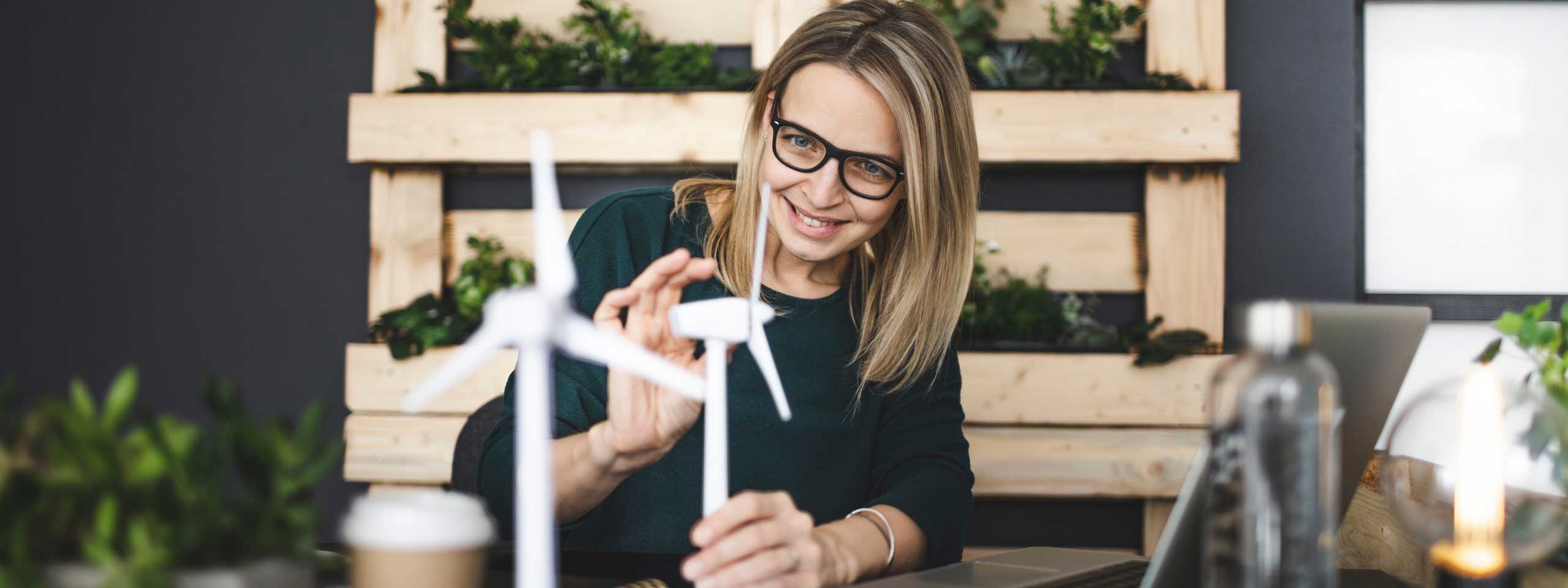 Une femme travaille sur un modèle d'éolienne à un bureau avec des plantes et un ordinateur portable dans un bureau moderne.