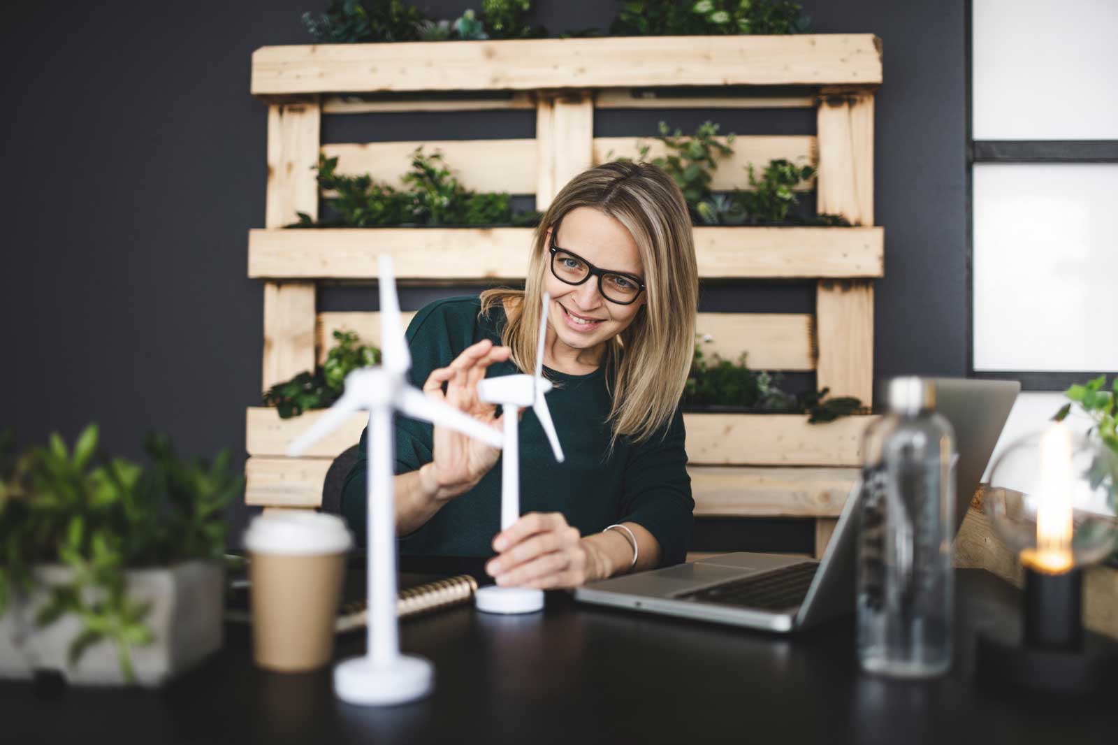 Une femme travaille à un bureau avec des éoliennes miniatures, un ordinateur portable et des plantes en arrière-plan.