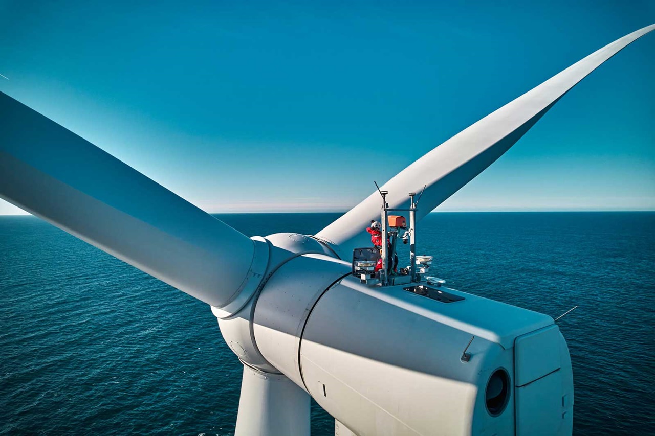 Un technicien travaille au sommet d'une éolienne surplombant l'océan sous un ciel bleu clair.