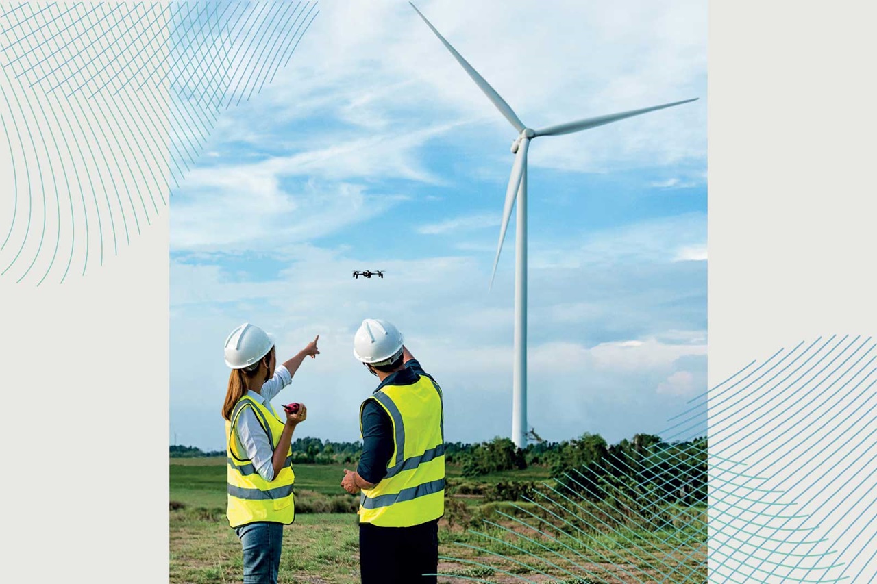 Deux professionnels en gilets de sécurité regardent un drone en vol près d'une éolienne sous un ciel bleu.