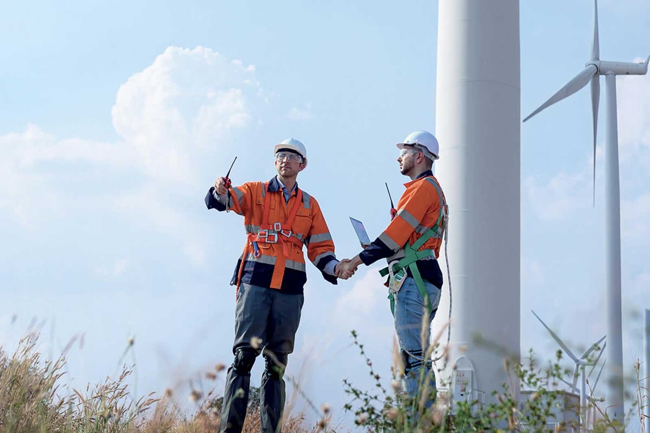 Deux travailleurs en équipement de sécurité discutent à côté d'une éolienne, tenant une tablette et un dispositif de communication, avec un ciel bleu en arrière-plan.