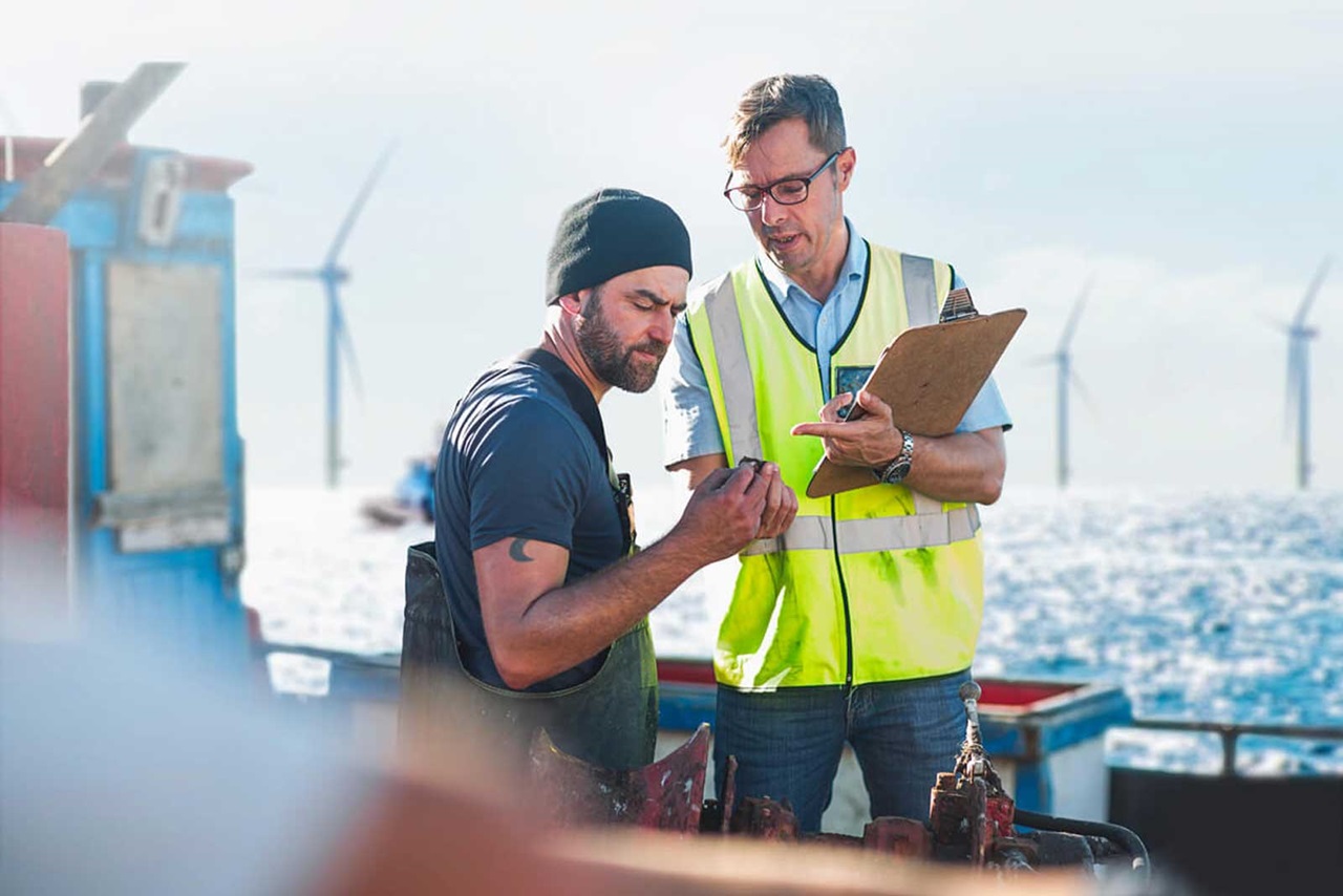 Deux hommes discutent sur un bateau, avec des éoliennes visibles en arrière-plan sous un ciel dégagé.