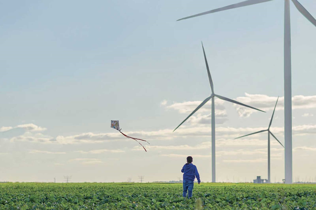 Un enfant fait voler un cerf-volant dans un champ, avec des éoliennes en arrière-plan sous un ciel dégagé.
