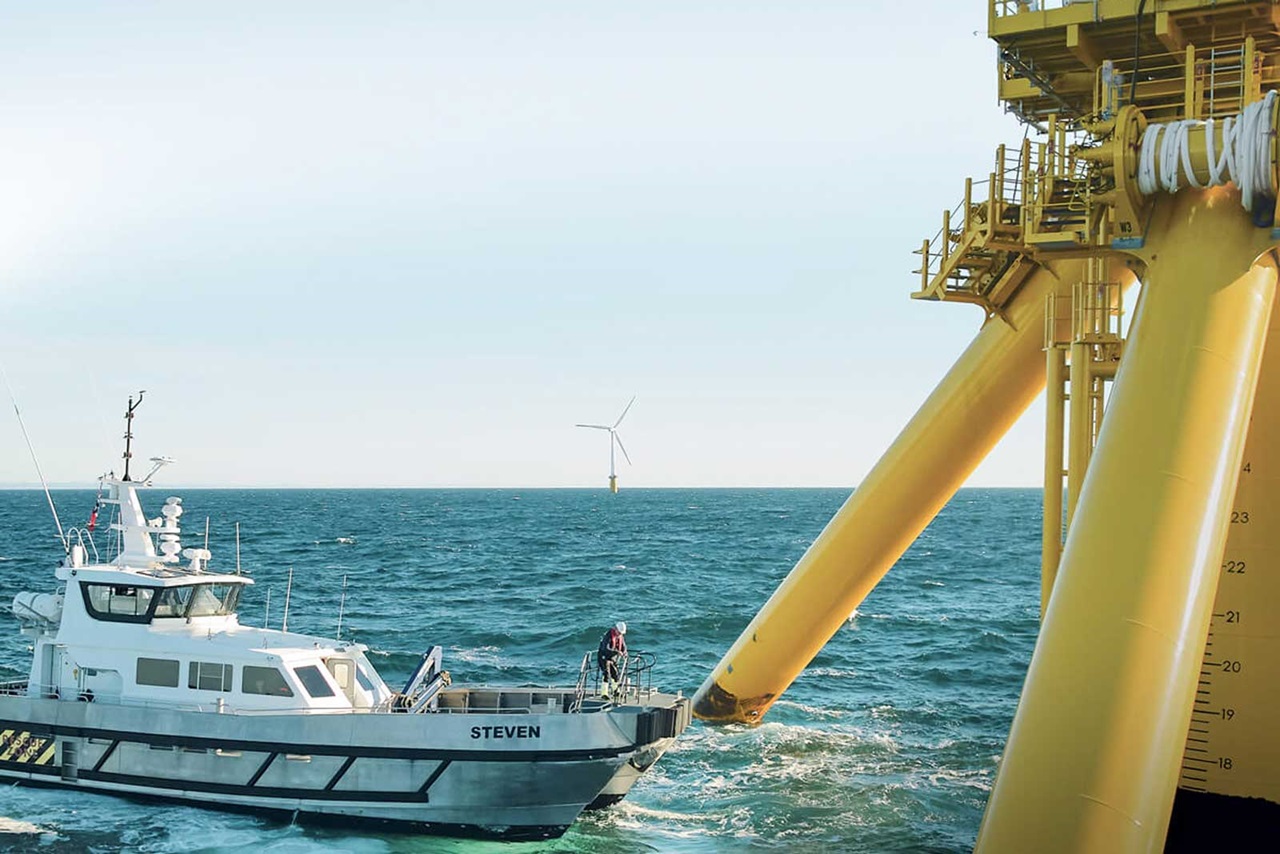 Un navire nommé 'Steven' s'approche d'une éolienne en mer, avec des structures de soutien jaunes et une mer calme en arrière-plan.