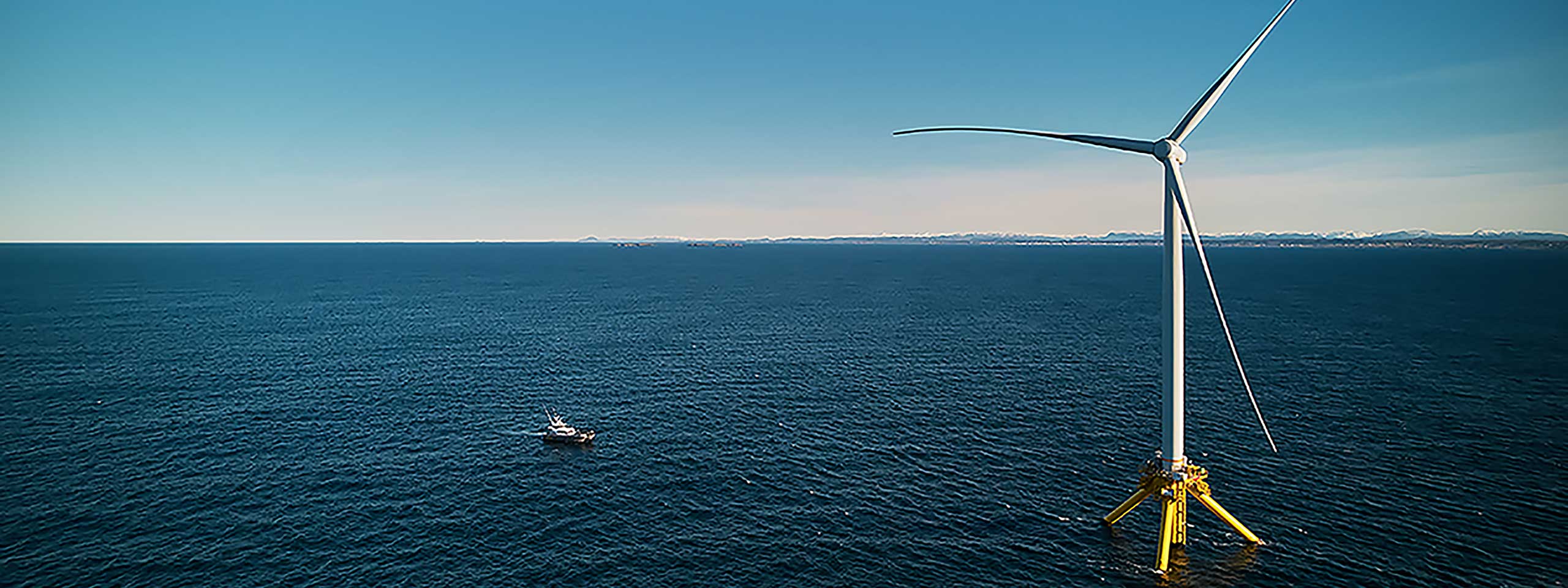 Une grande éolienne se dresse au large, avec un bateau à proximité et un ciel bleu clair au-dessus.