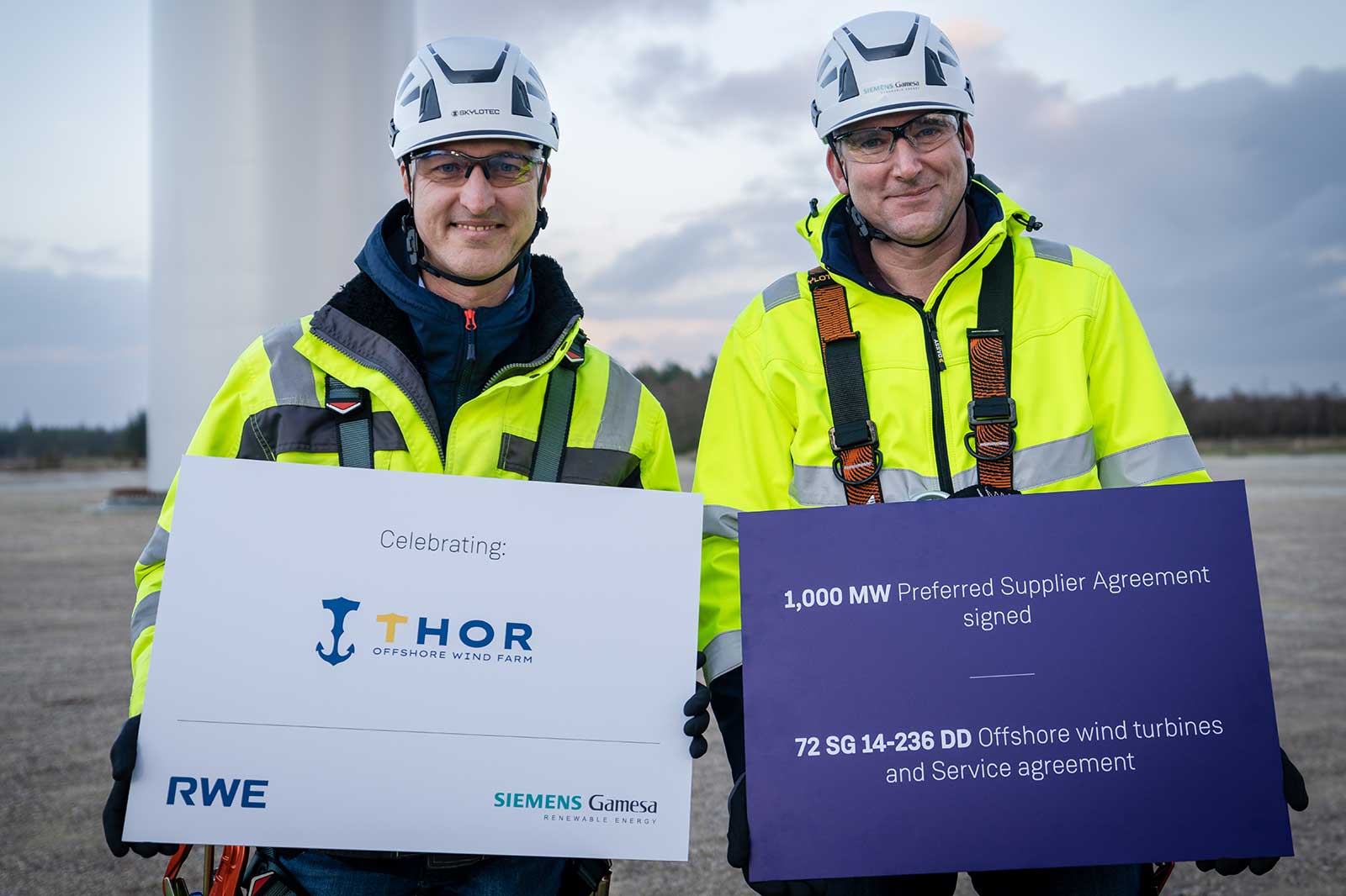 Two individuals in safety gear celebrate the THOR Offshore Wind Farm agreement, holding signs with contract details.
