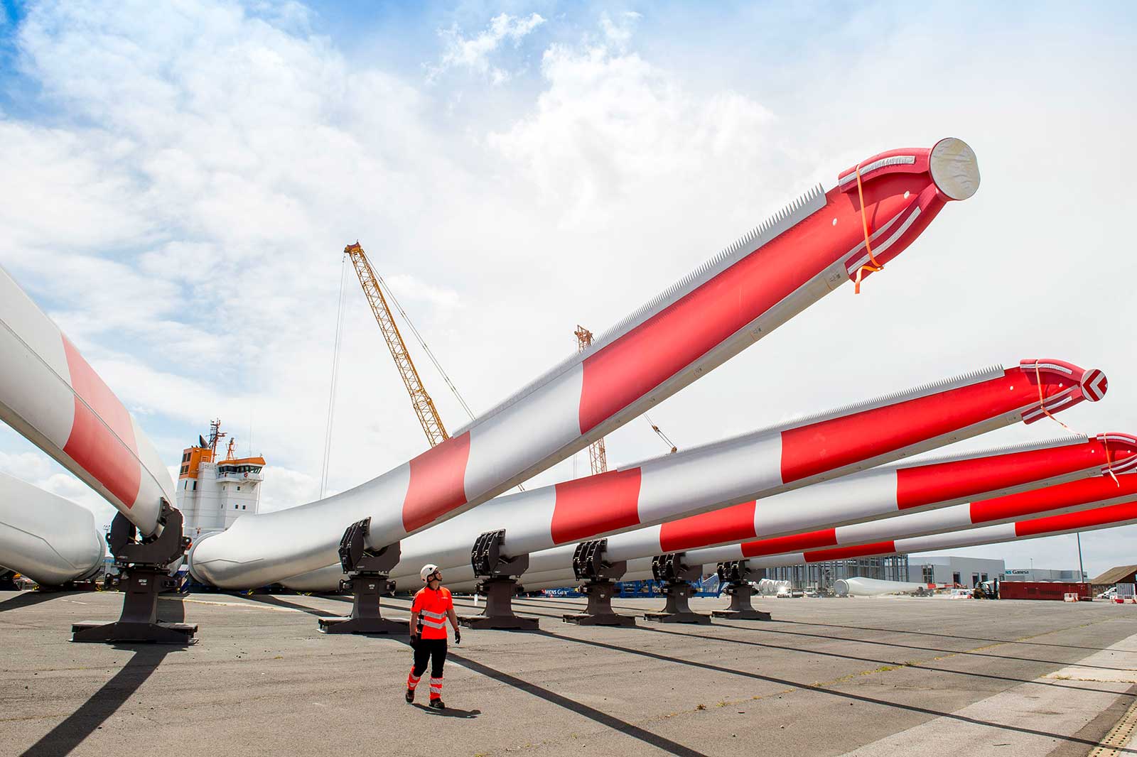 A worker in a safety helmet walks past large wind turbine blades, marked with red and white stripes, at a construction site.