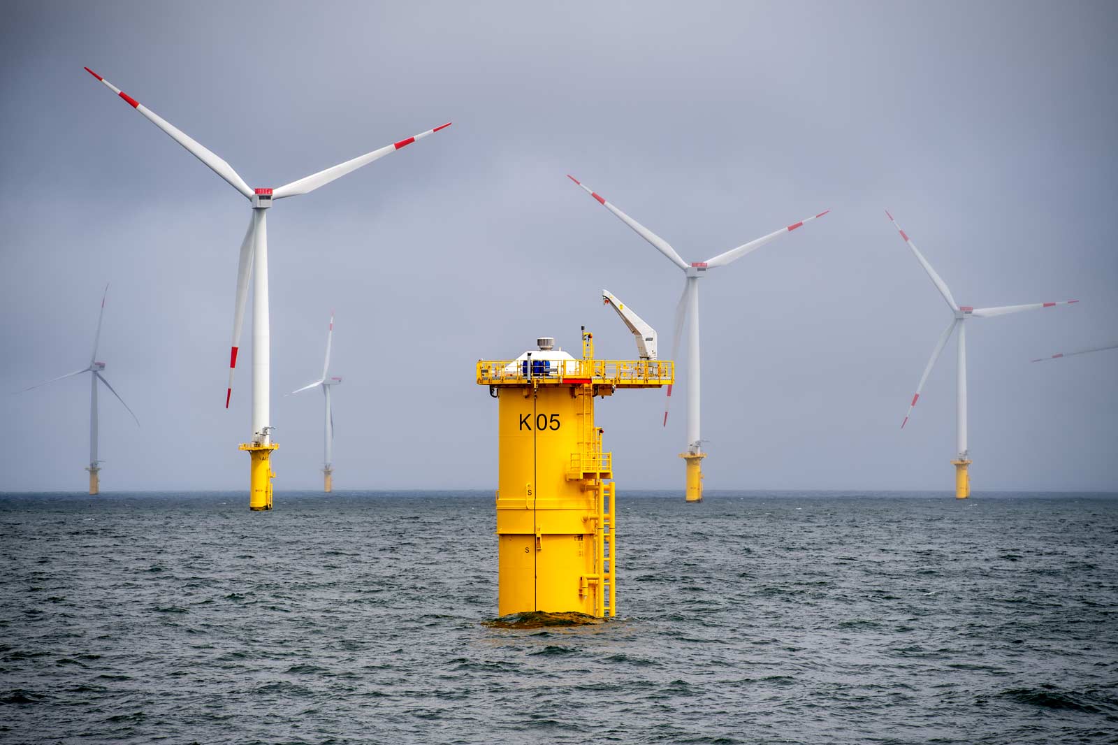 A bright yellow offshore wind turbine platform stands in the sea, surrounded by several white turbines under overcast skies.