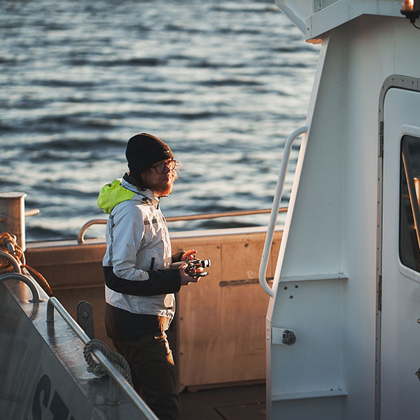 Une personne se tient sur un bateau, portant une veste avec une capuche lumineuse, tenant une télécommande avec de l'eau en arrière-plan.
