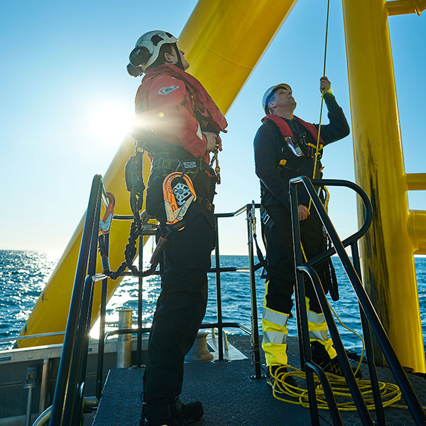 Deux travailleurs en équipements de sécurité manipulent des équipements sur une plateforme offshore, avec la mer et le soleil en arrière-plan.
