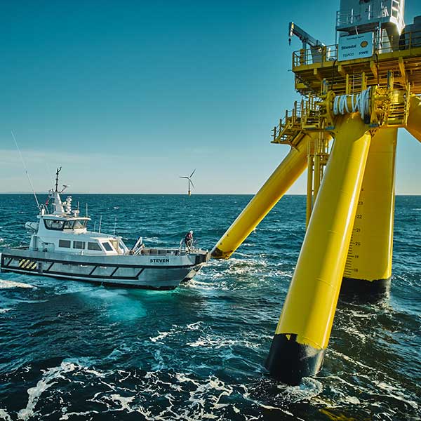 Un bateau nommé Steven s'approche d'une grande plateforme offshore jaune avec une éolienne en arrière-plan sous un ciel bleu clair.