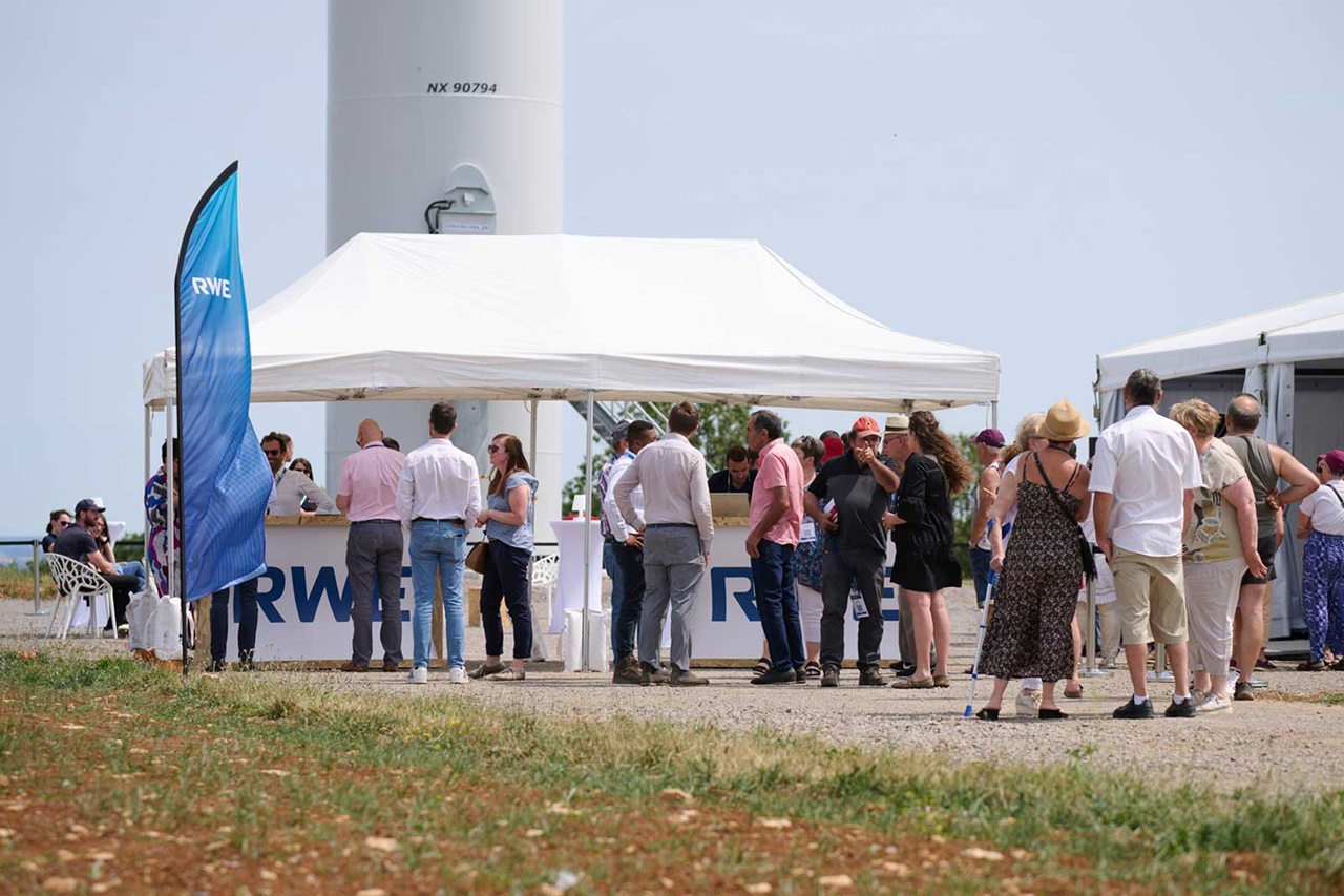 Un groupe de personnes se rassemble autour d'une tente avec une bannière RWE, près d'une éolienne.