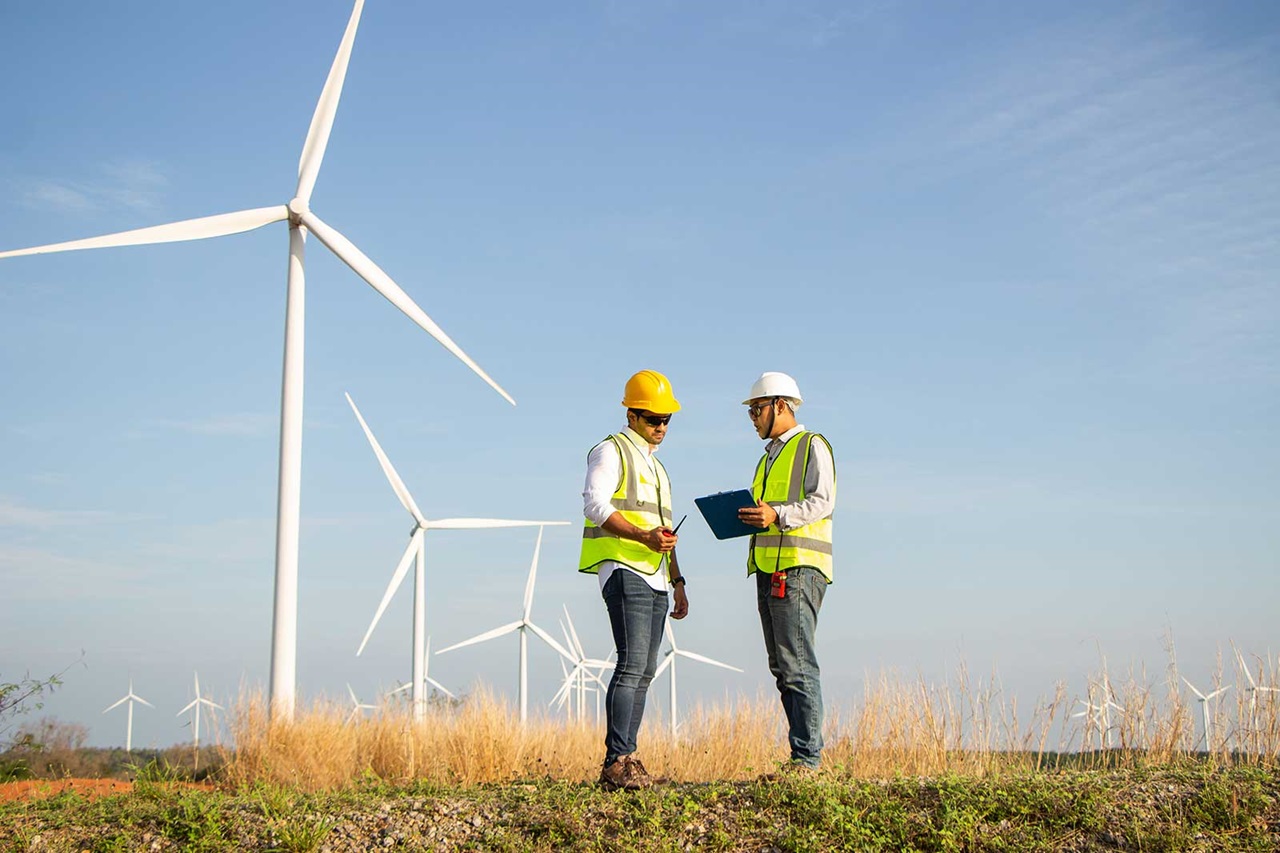Deux travailleurs en gilets de sécurité et casques discutent des plans près des éoliennes sous un ciel bleu clair.