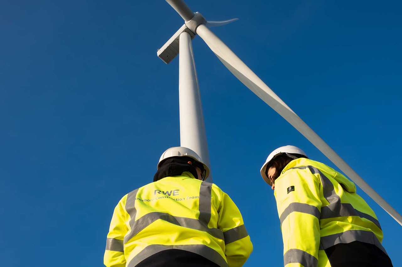 Two workers in safety equipment inspect a large wind turbine against a clear blue sky.