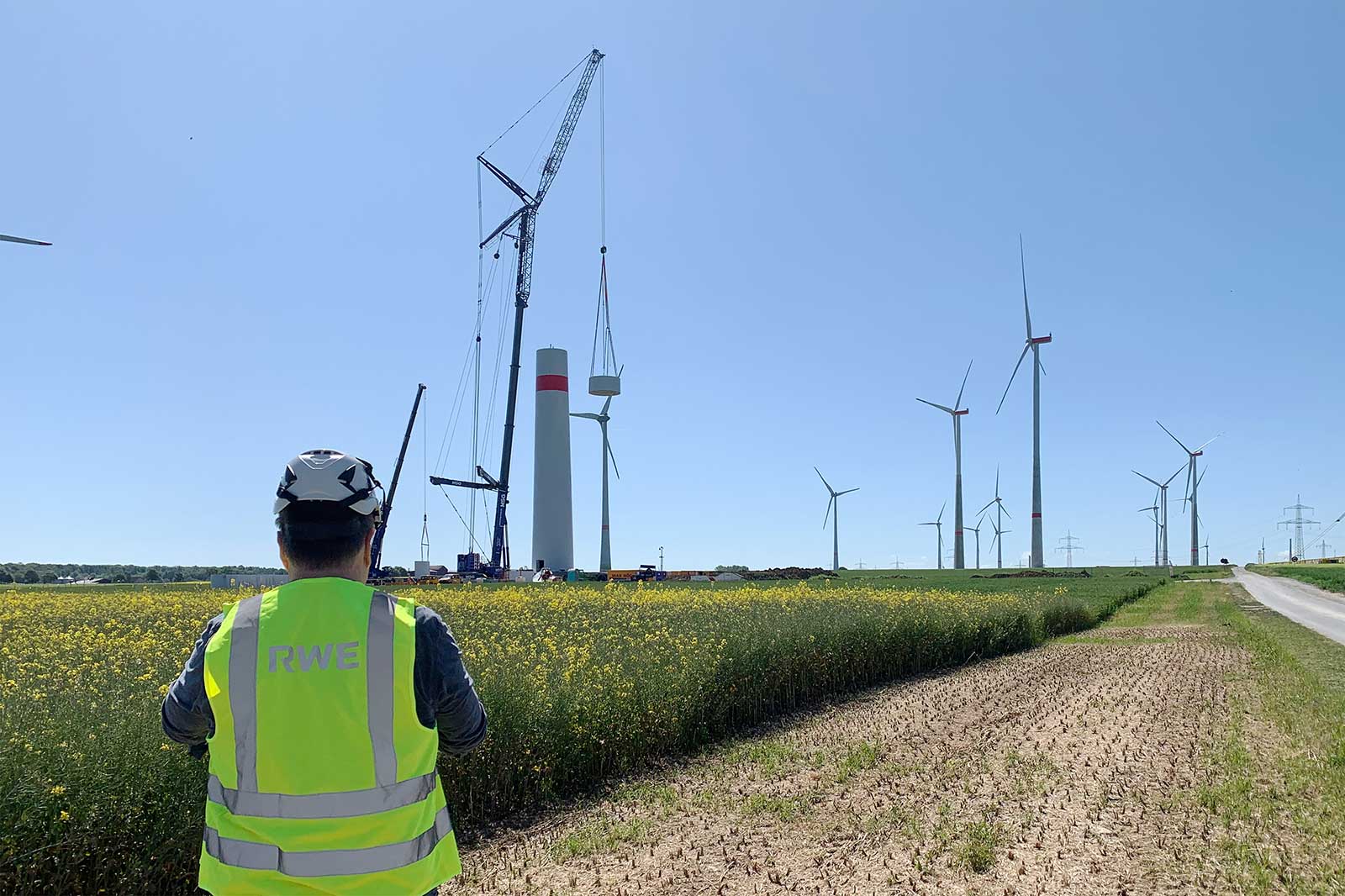 Un travailleur en gilet de haute visibilité observe des éoliennes et de l'équipement de construction dans un champ sous un ciel bleu clair.