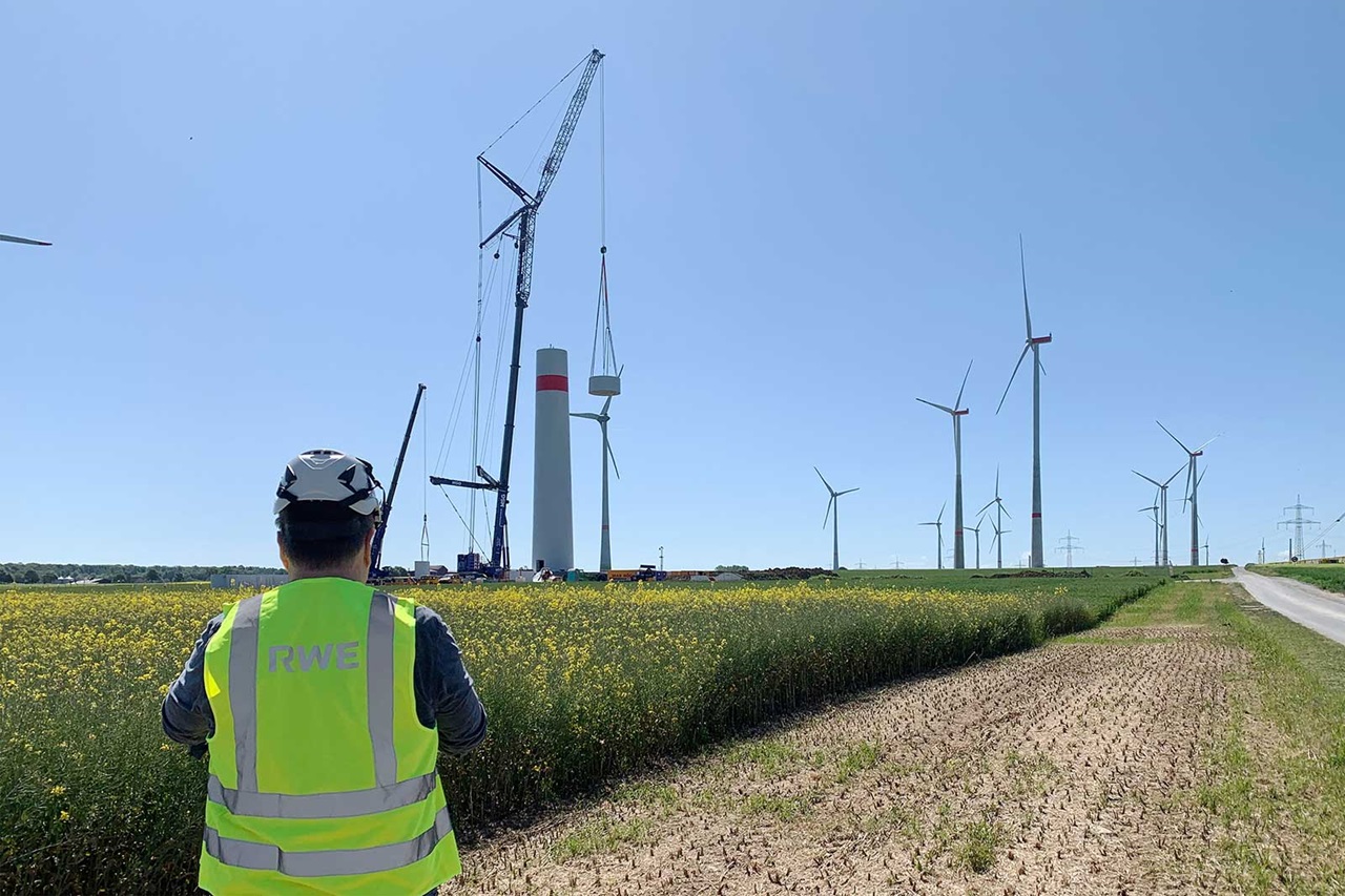 A worker in a high-visibility vest observes wind turbines and construction equipment in a field under a clear blue sky.