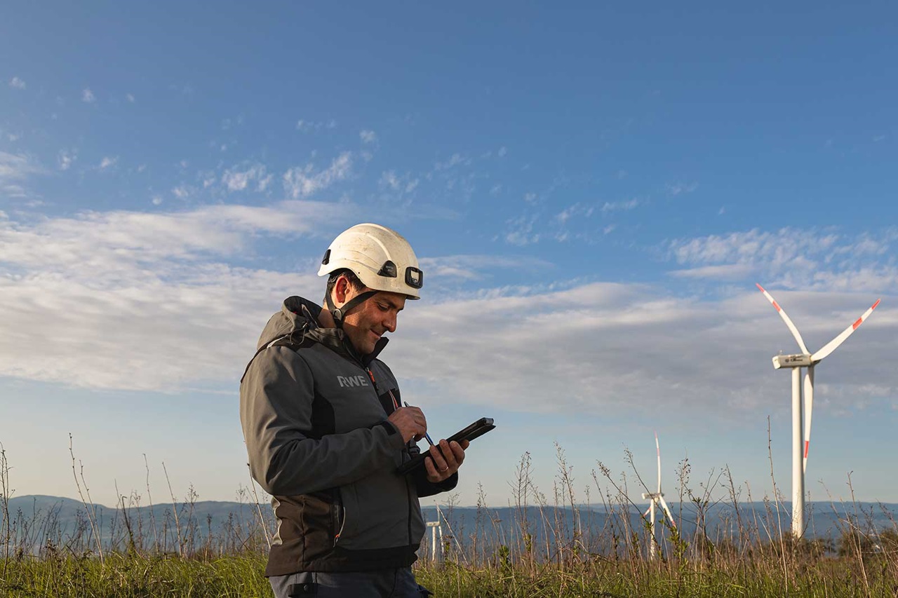 A worker wearing a helmet is using a device outside near wind turbines under a blue sky.
