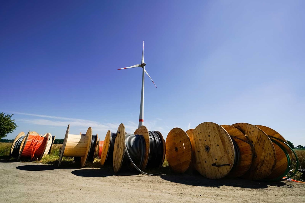 A row of cable reels in the foreground with a wind turbine in the background under a light blue sky.