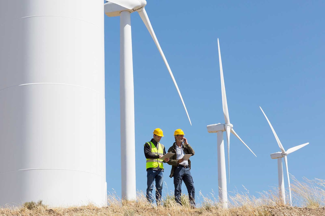 Two workers in safety clothing stand near wind turbines, examining documents under a clear blue sky.