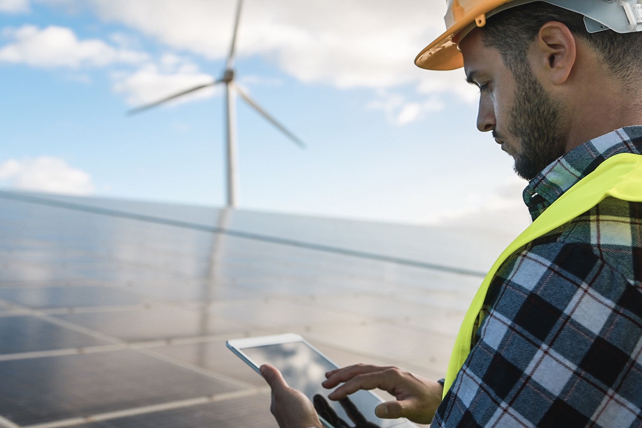 Un travailleur en casque de protection et gilet haute visibilité utilise une tablette, avec des panneaux solaires et une éolienne en arrière-plan.
