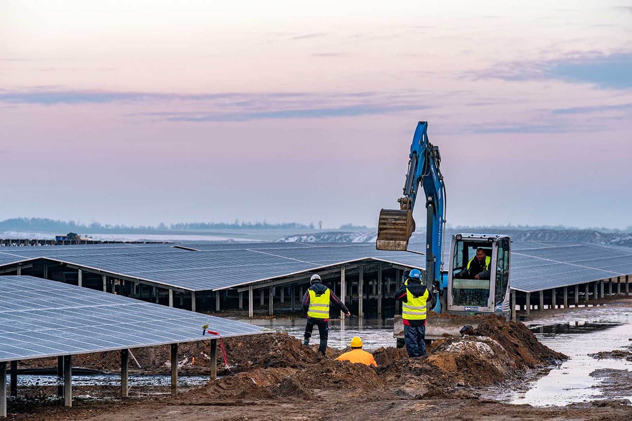 Des ouvriers de construction utilisent des machines sur un parc solaire, avec des rangées de panneaux solaires et un ciel rosé en arrière-plan.