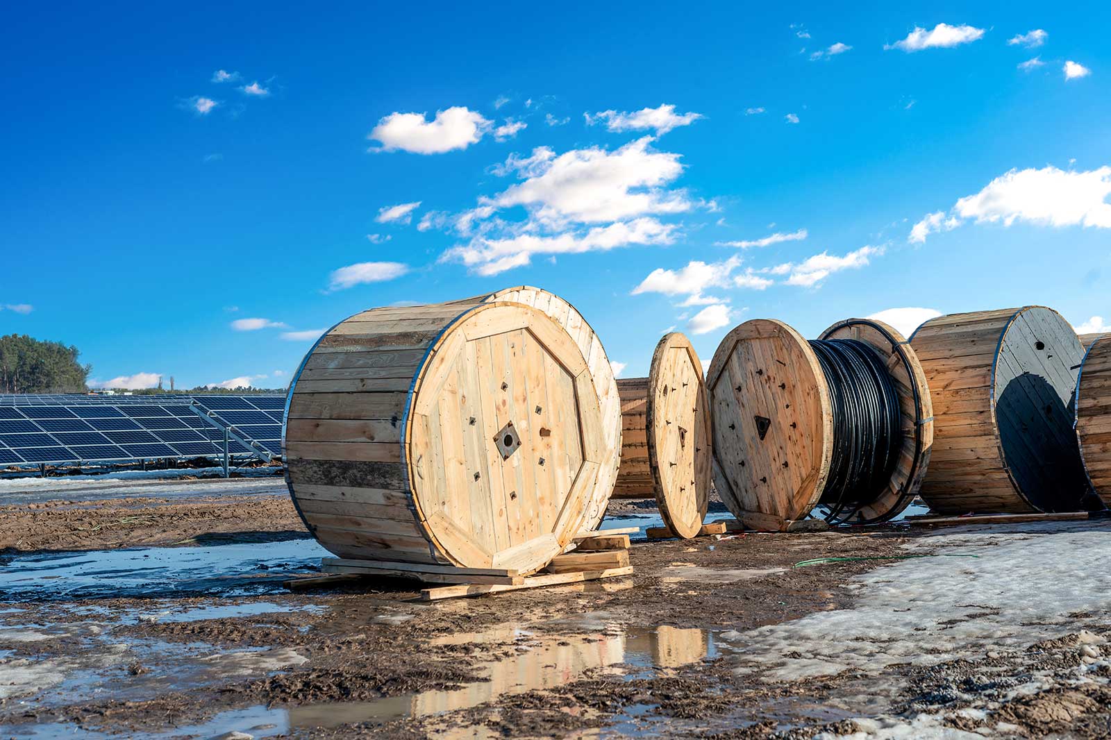 Several wooden cable reels are placed on muddy ground, with solar panels and a clear blue sky in the background.