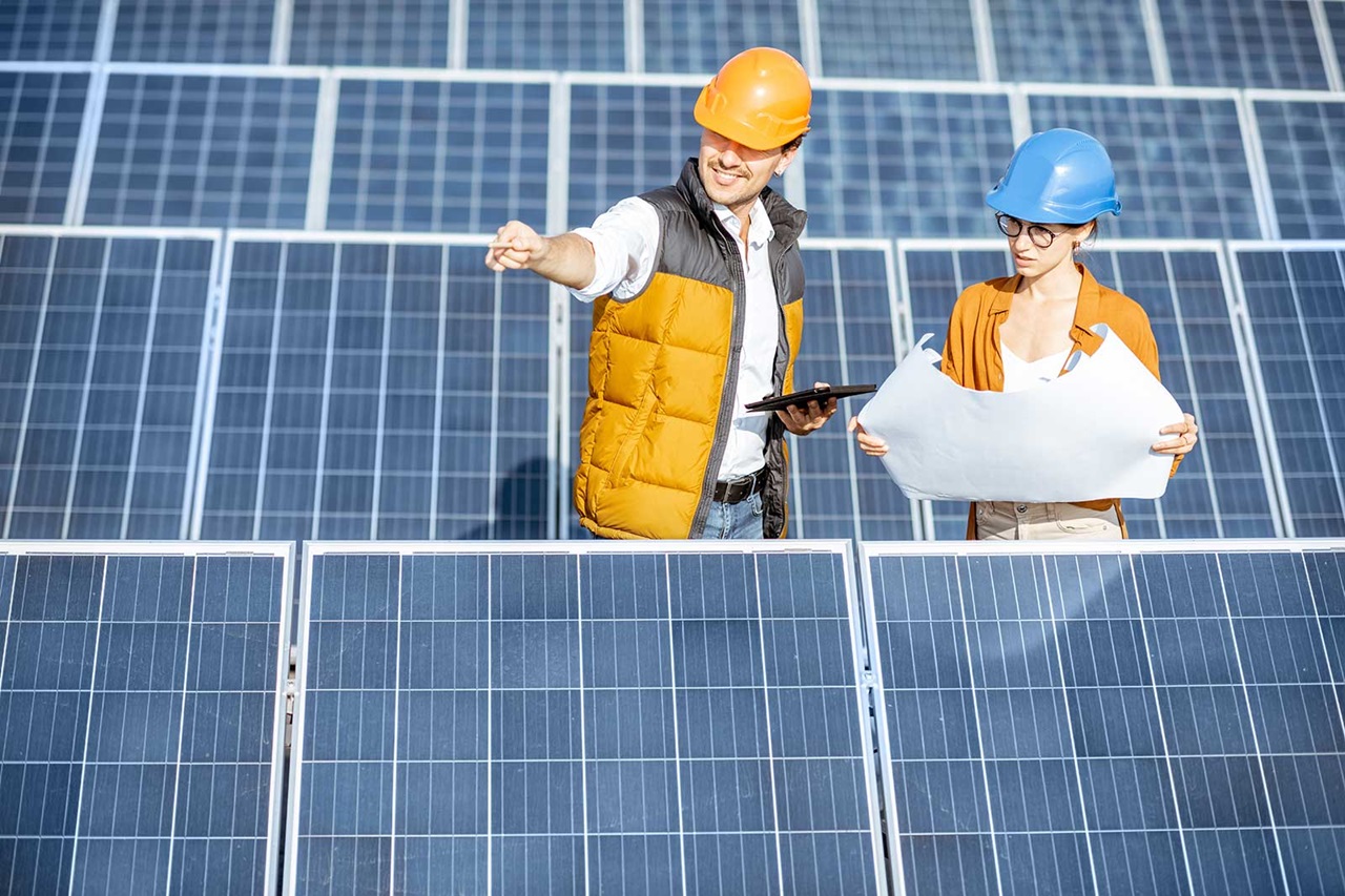 Deux travailleurs de la construction discutent des plans devant des panneaux solaires, l'un tenant une tablette et l'autre un plan.