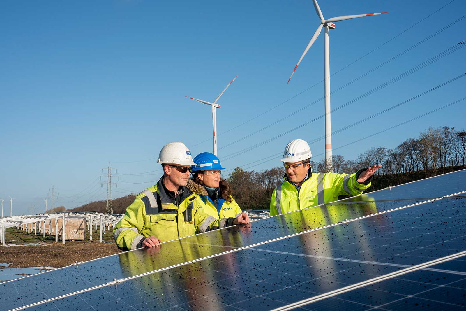 Three workers in helmets examine solar panels under a clear blue sky, with wind turbines in the background.