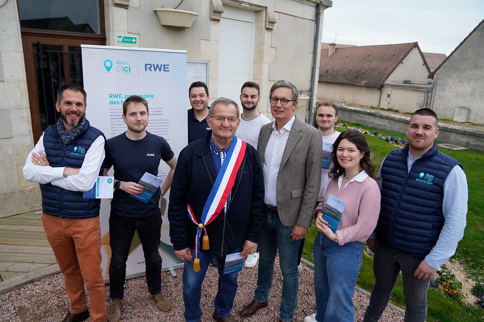 Groupe de personnes posant à l'extérieur avec une bannière pour RWE. Le maire est au centre avec une écharpe.