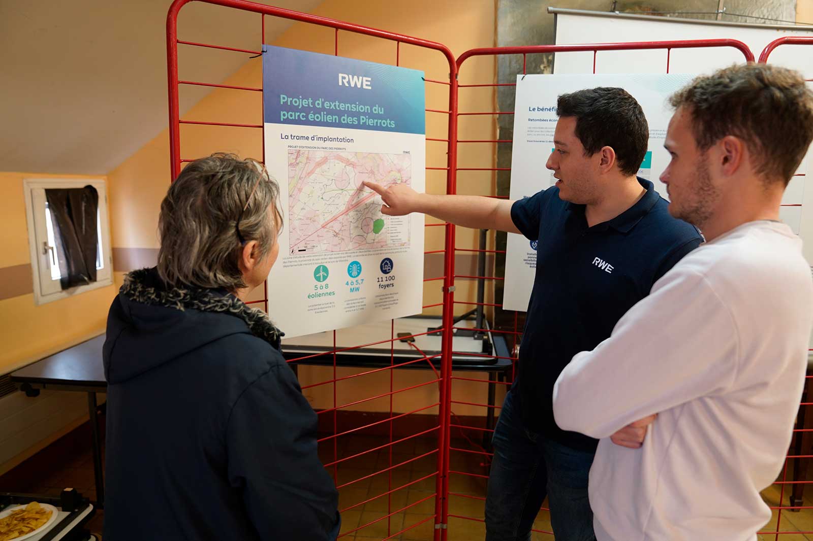 Un groupe discute d'un projet de parc éolien, montrant une carte et des affiches d'information lors d'une réunion communautaire.