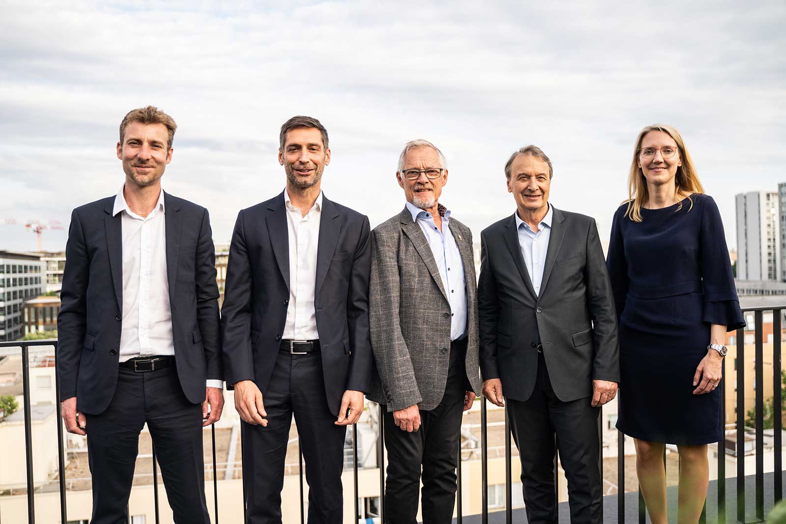 Cinq personnes debout en tenue soignée sur un balcon avec une vue sur la ville, sous un ciel partiellement nuageux.