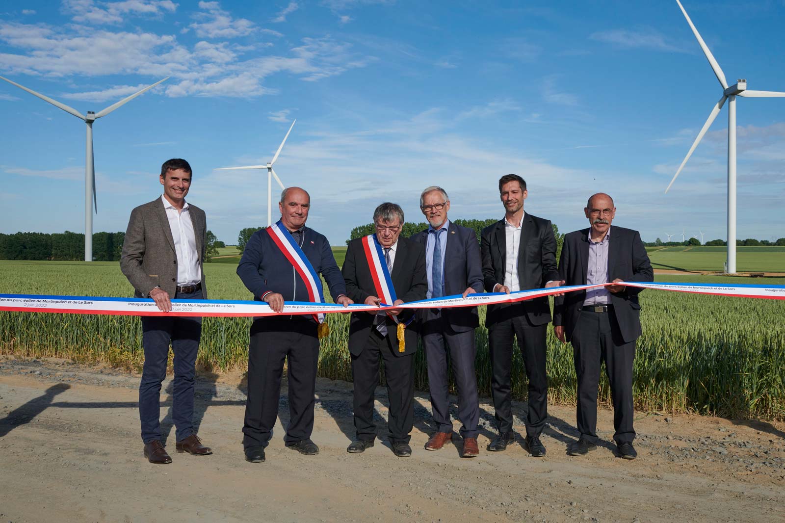 Un groupe d'officiels lors d'une cérémonie d'inauguration pour un parc éolien, avec des éoliennes et des champs verdoyants en arrière-plan.