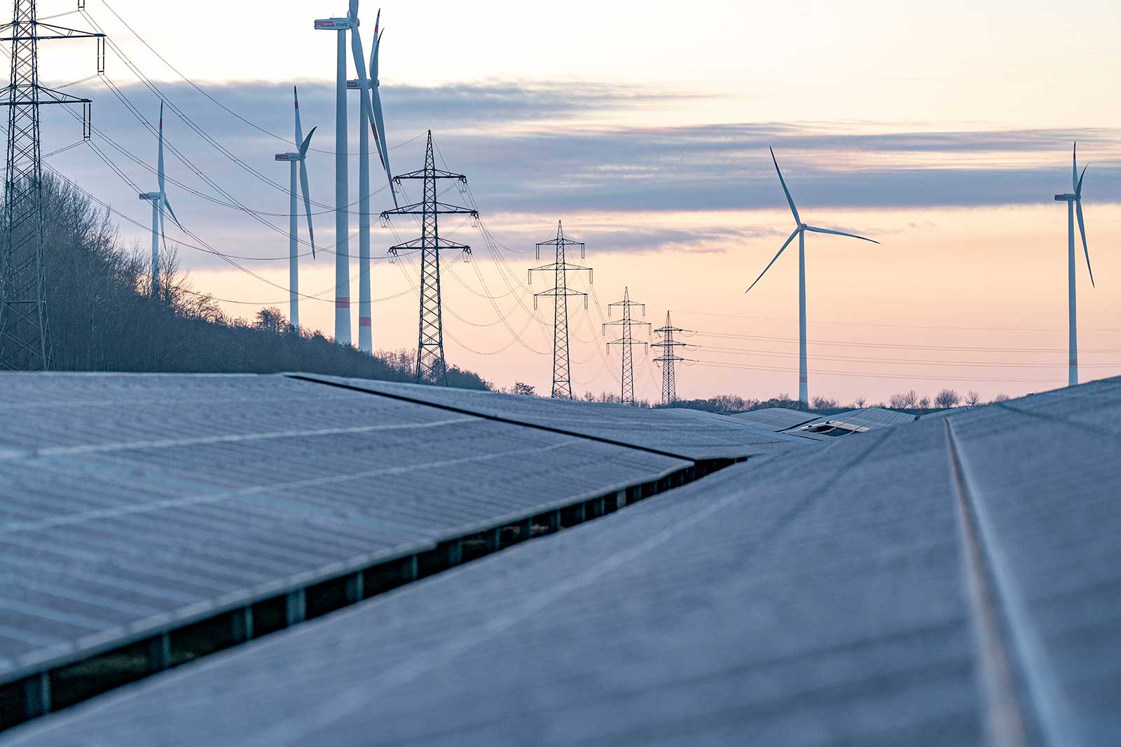 Un paysage avec des panneaux solaires au premier plan, des éoliennes en arrière-plan et des lignes électriques sous un ciel pastel.