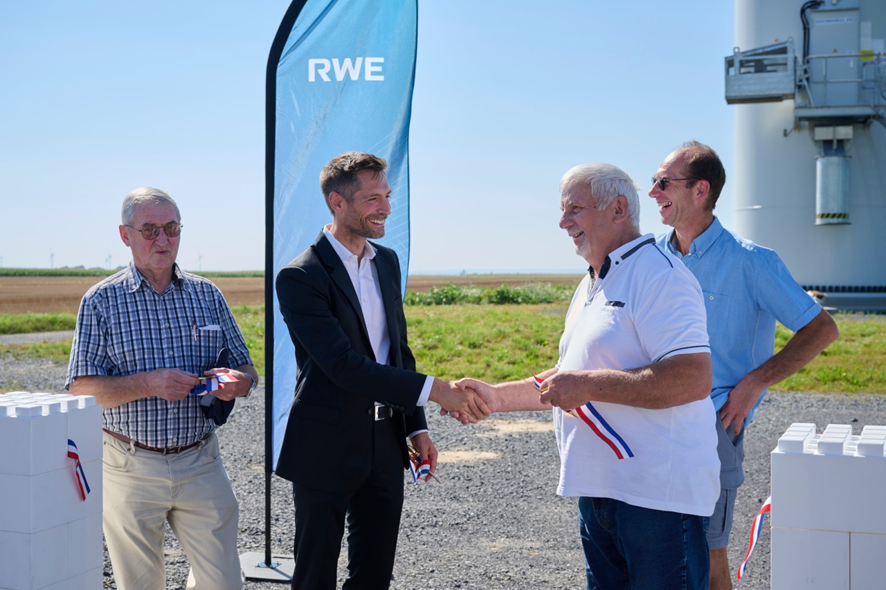 Four individuals engaged in a handshake at an outdoor event, with a blue RWE banner in the background and white structures nearby.