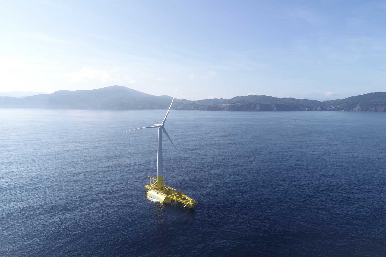 A floating wind turbine stands on calm waters with mountains in the background under a clear sky.