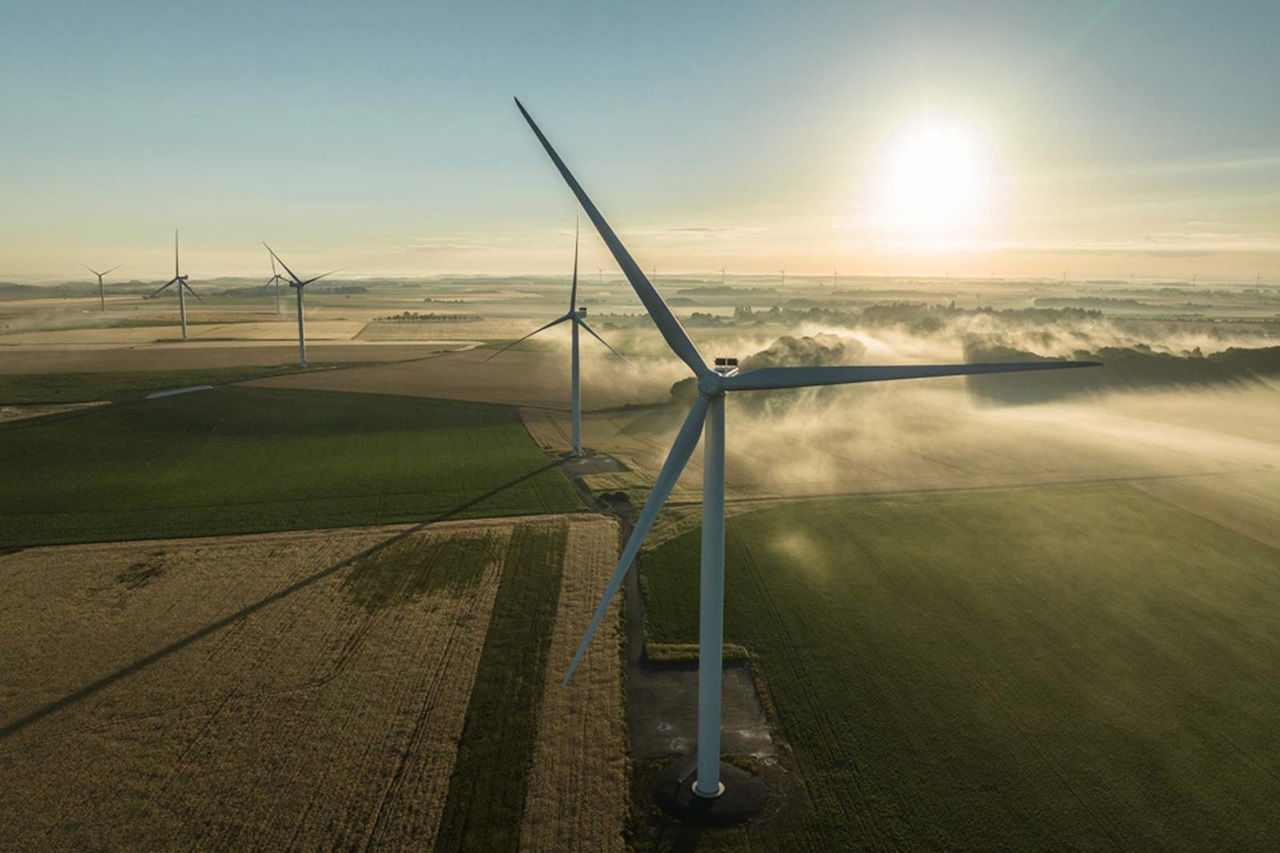Vue aérienne de turbines éoliennes dans un paysage verdoyant au lever du soleil, avec du brouillard s'élevant doucement sur les champs.