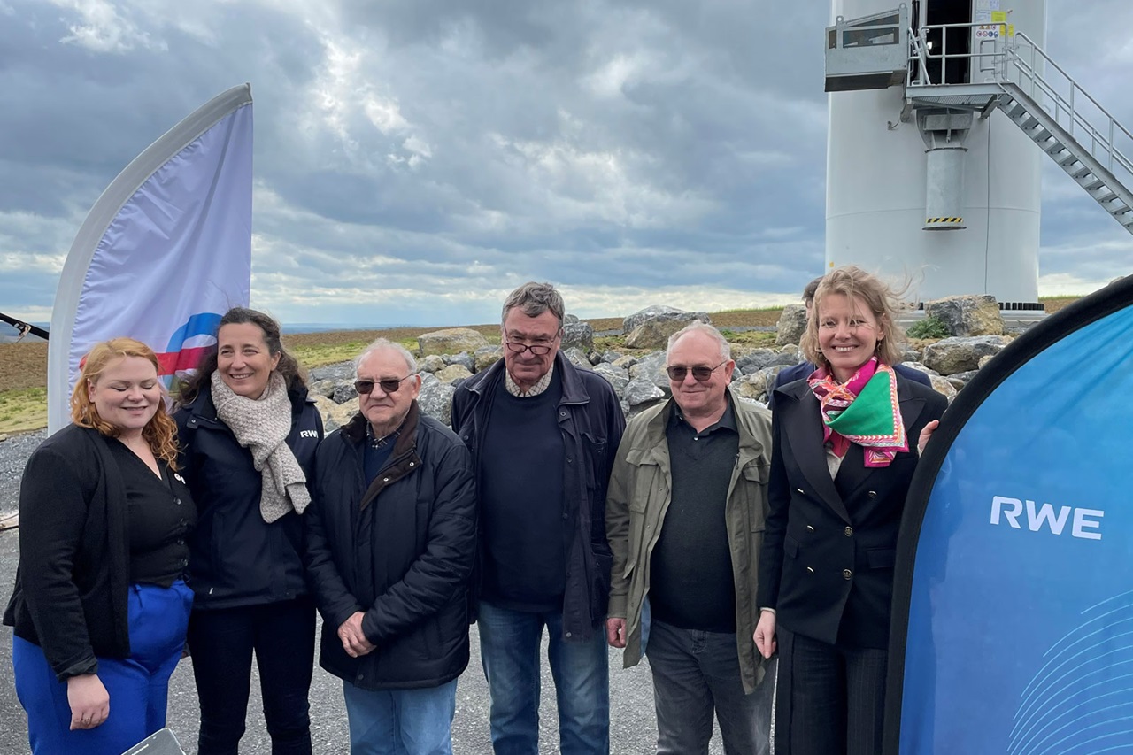Groupe de personnes posant à l'extérieur près d'une éolienne, avec un drapeau et une tente bleue en arrière-plan, sous un ciel nuageux.