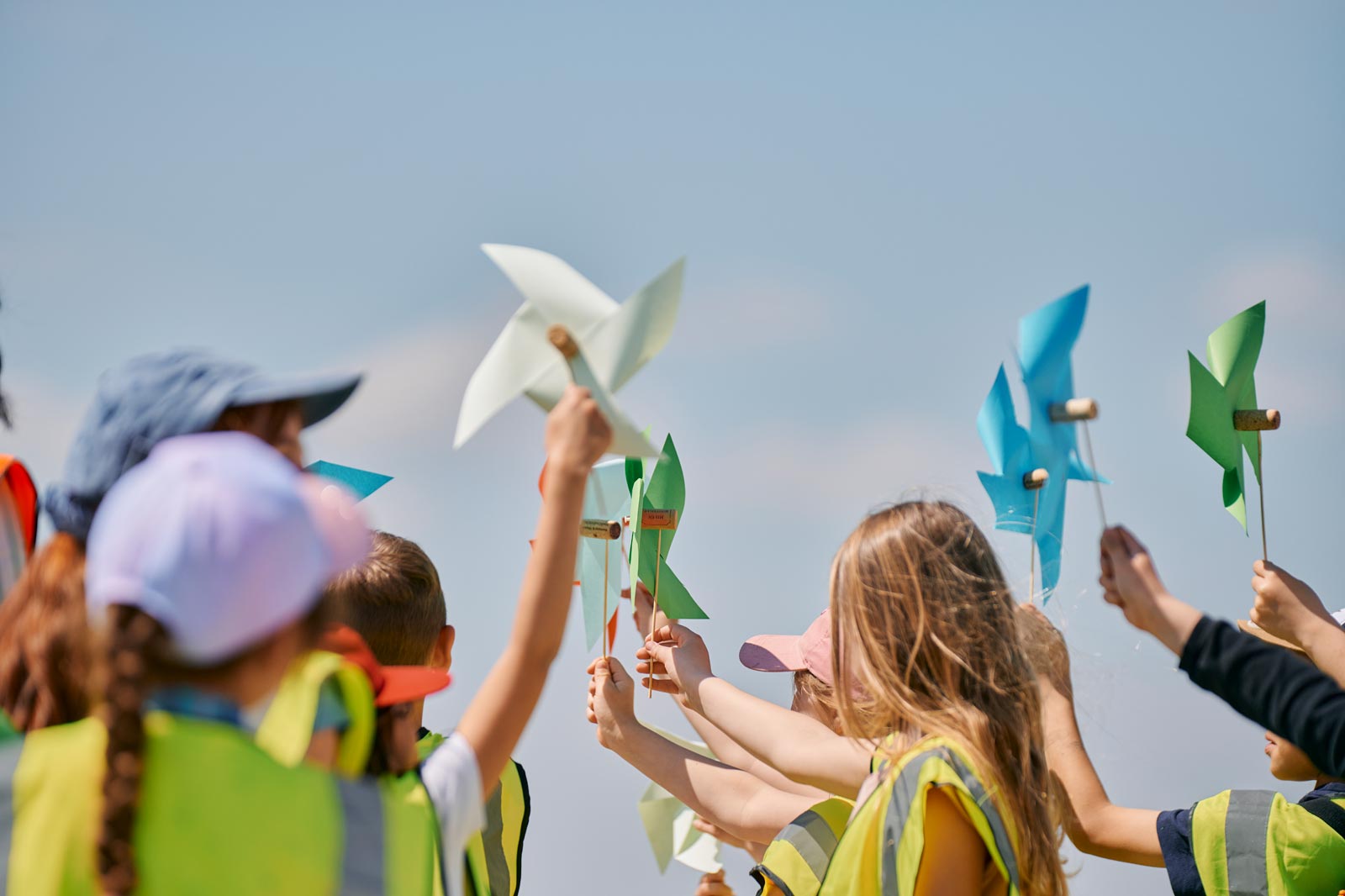 Children in brightly coloured vests joyfully wave handmade pinwheels under a clear blue sky.