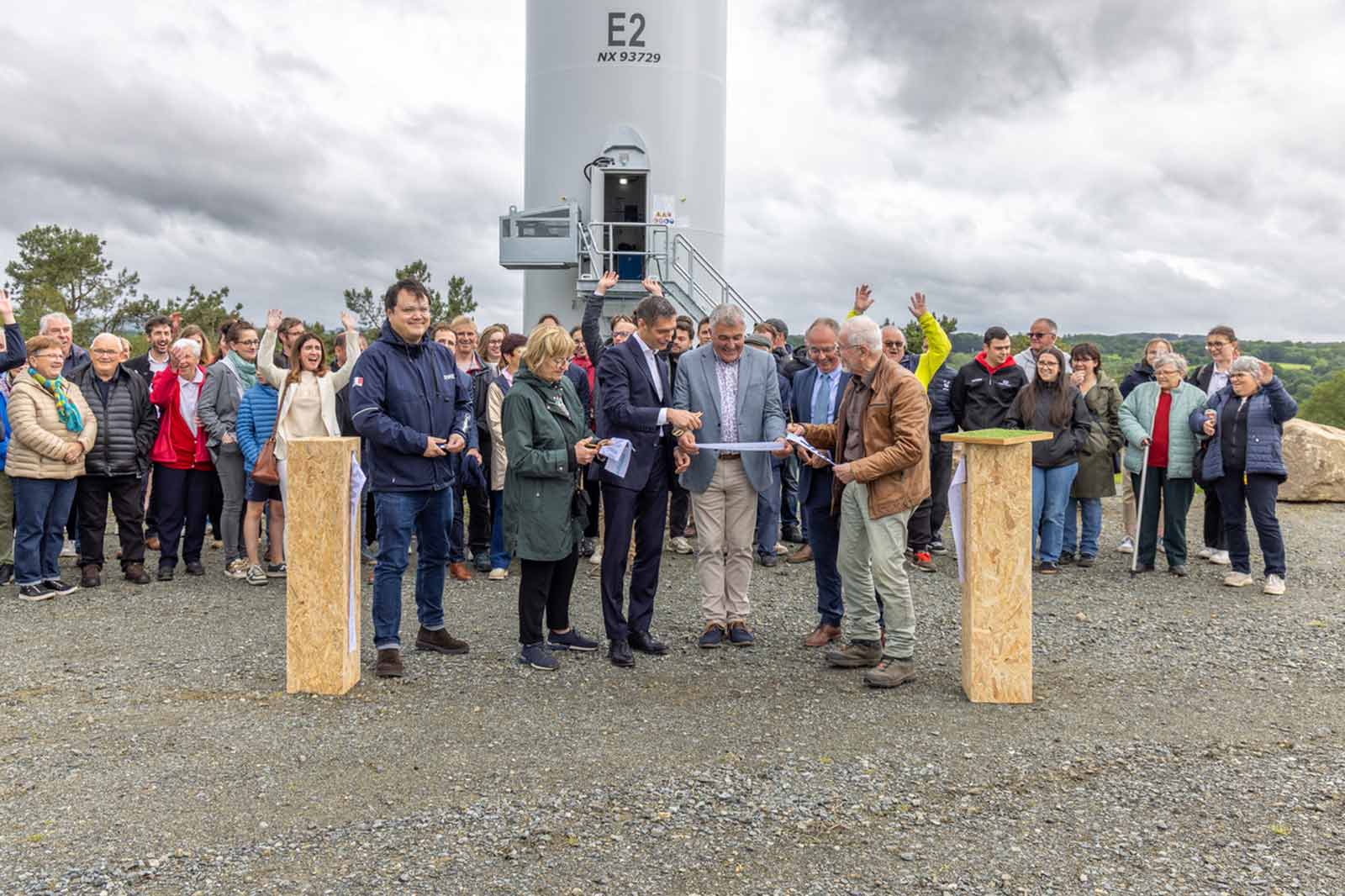 Une foule se rassemble sur un site d'éolienne, avec plusieurs personnes participant à une cérémonie d'inauguration sous un ciel nuageux.