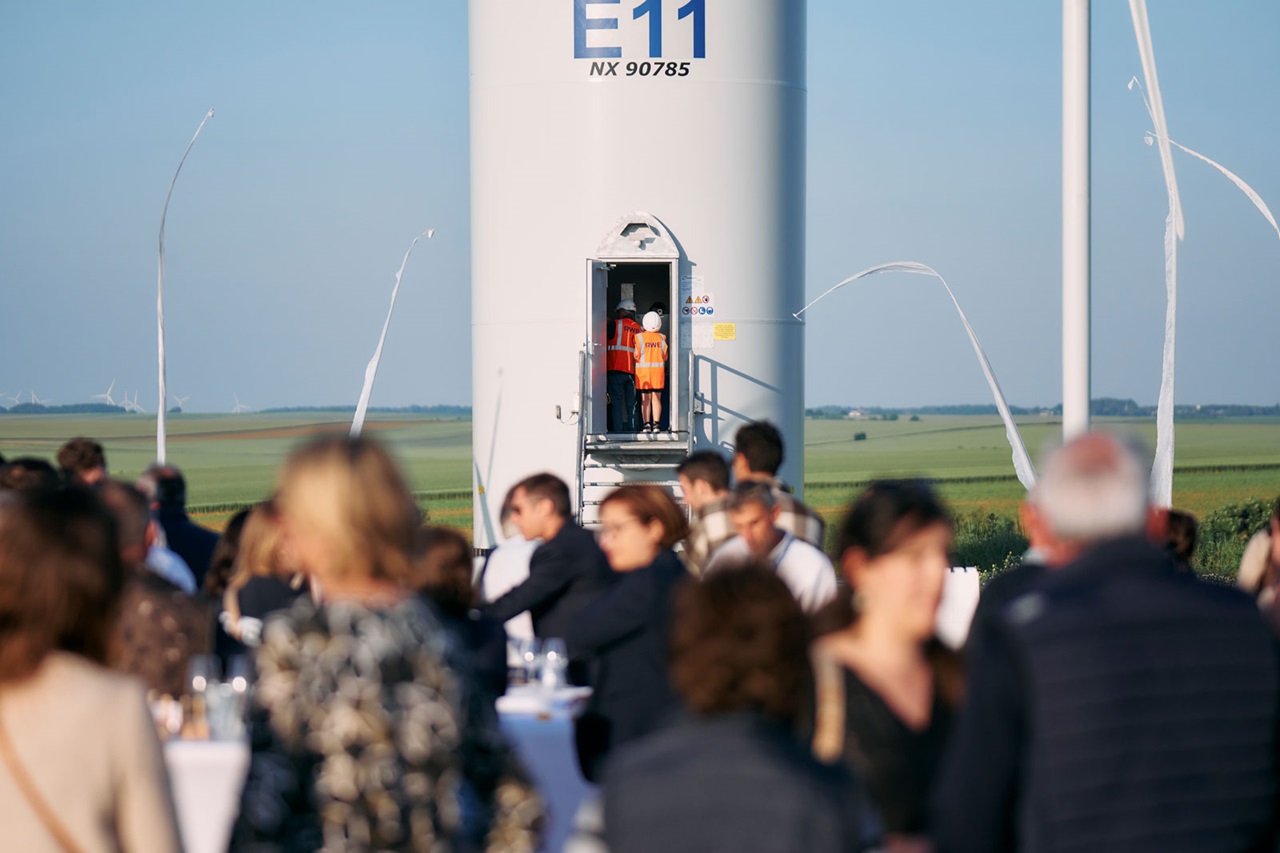 Une foule se rassemble près d'une éolienne avec deux travailleurs se préparant à monter. Le paysage présente des champs verts et un ciel dégagé.