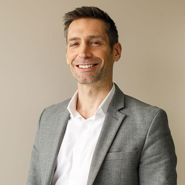 Un homme en blazer gris et chemise blanche se tient contre un fond neutre.