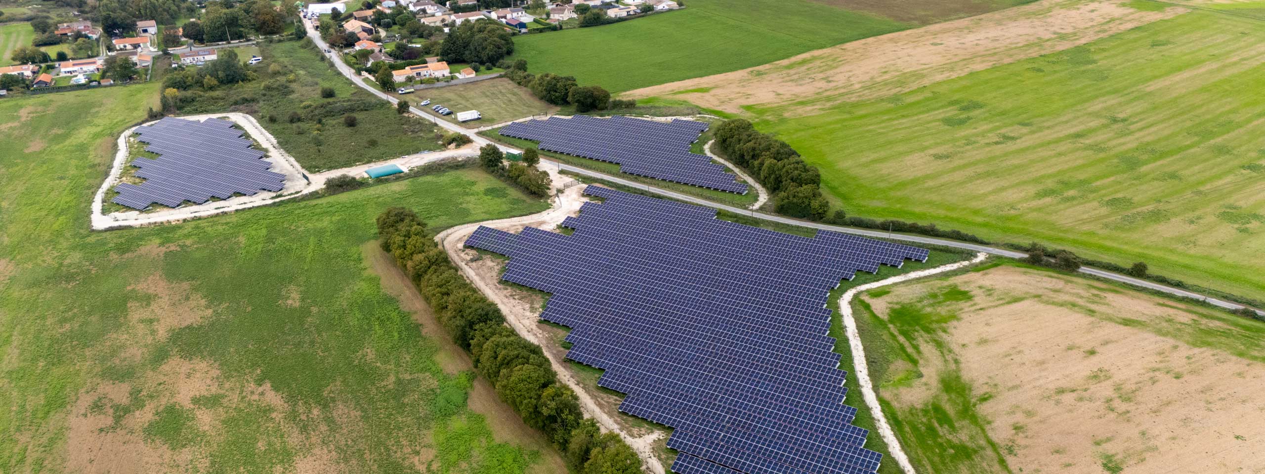 Vue aérienne de panneaux solaires disposés dans des champs, entourés de verdure et de maisons en arrière-plan.