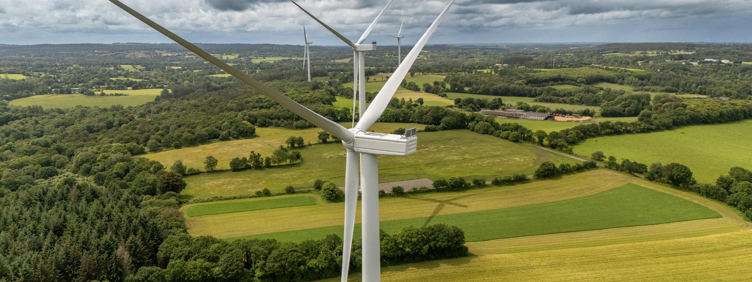 Vue aérienne d'éoliennes surplombant de luxuriants champs verts et des forêts sous un ciel nuageux.