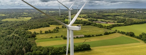 Vue aérienne d'éoliennes surplombant de luxuriants champs verts et des forêts sous un ciel nuageux.