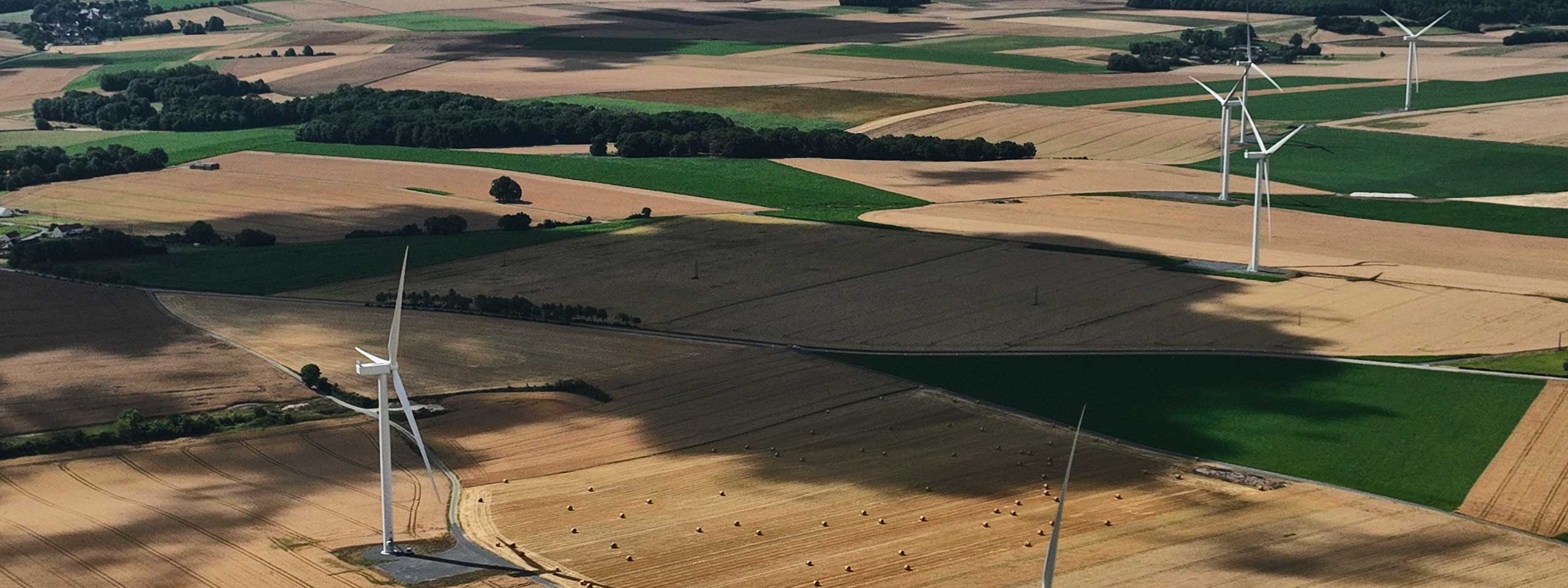 Vue aérienne d'un paysage avec des champs verts et bruns, parsemés d'éoliennes et de balles de foin.