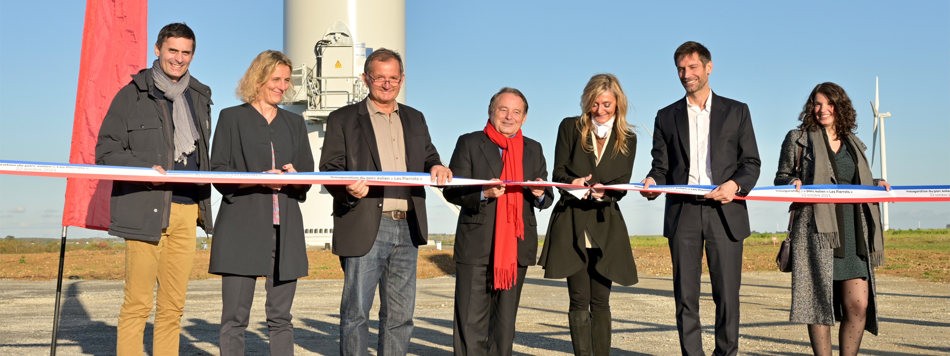 Un groupe de personnes coupe un ruban lors de l'inauguration d'un parc éolien, avec des éoliennes en arrière-plan.
