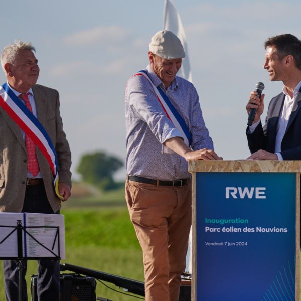 Un groupe d'officiels s'exprime lors de l'inauguration d'un parc éolien, avec un ciel bleu et des champs verts en arrière-plan.