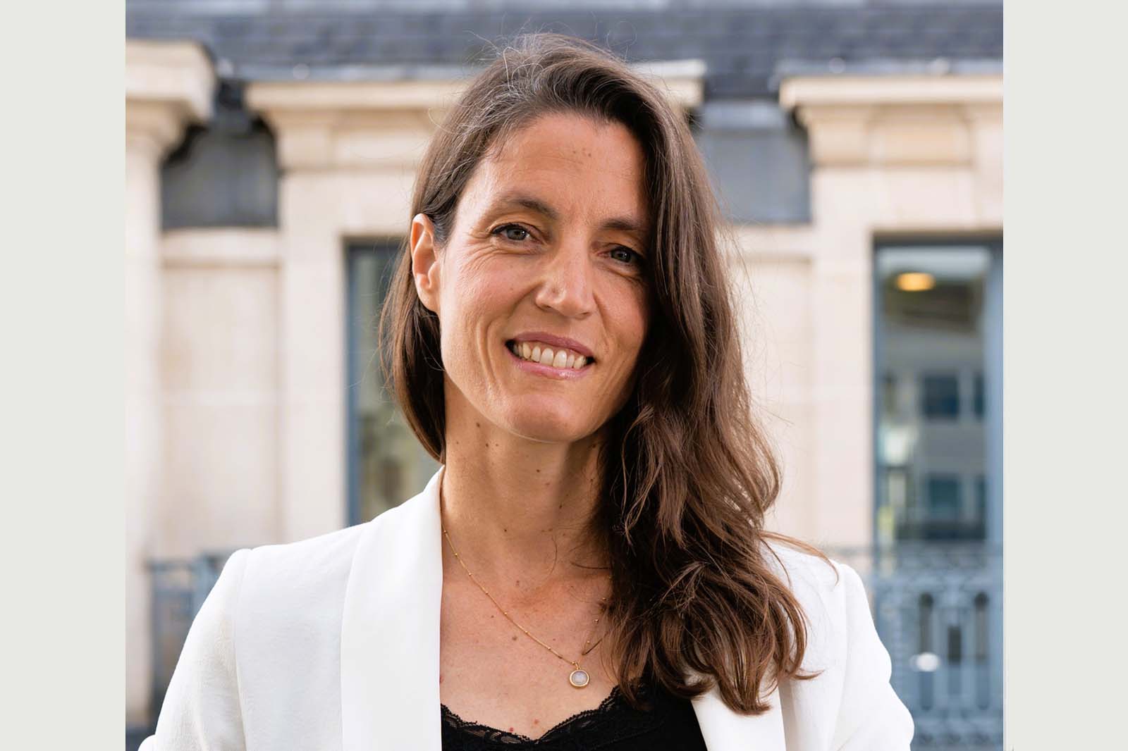 Close-up outdoor portrait of a woman with a blurred face, long brown hair over the shoulder, white blazer, black lace top, and gold necklace with a round pendant. Portrait of Gabrielle Dufour.